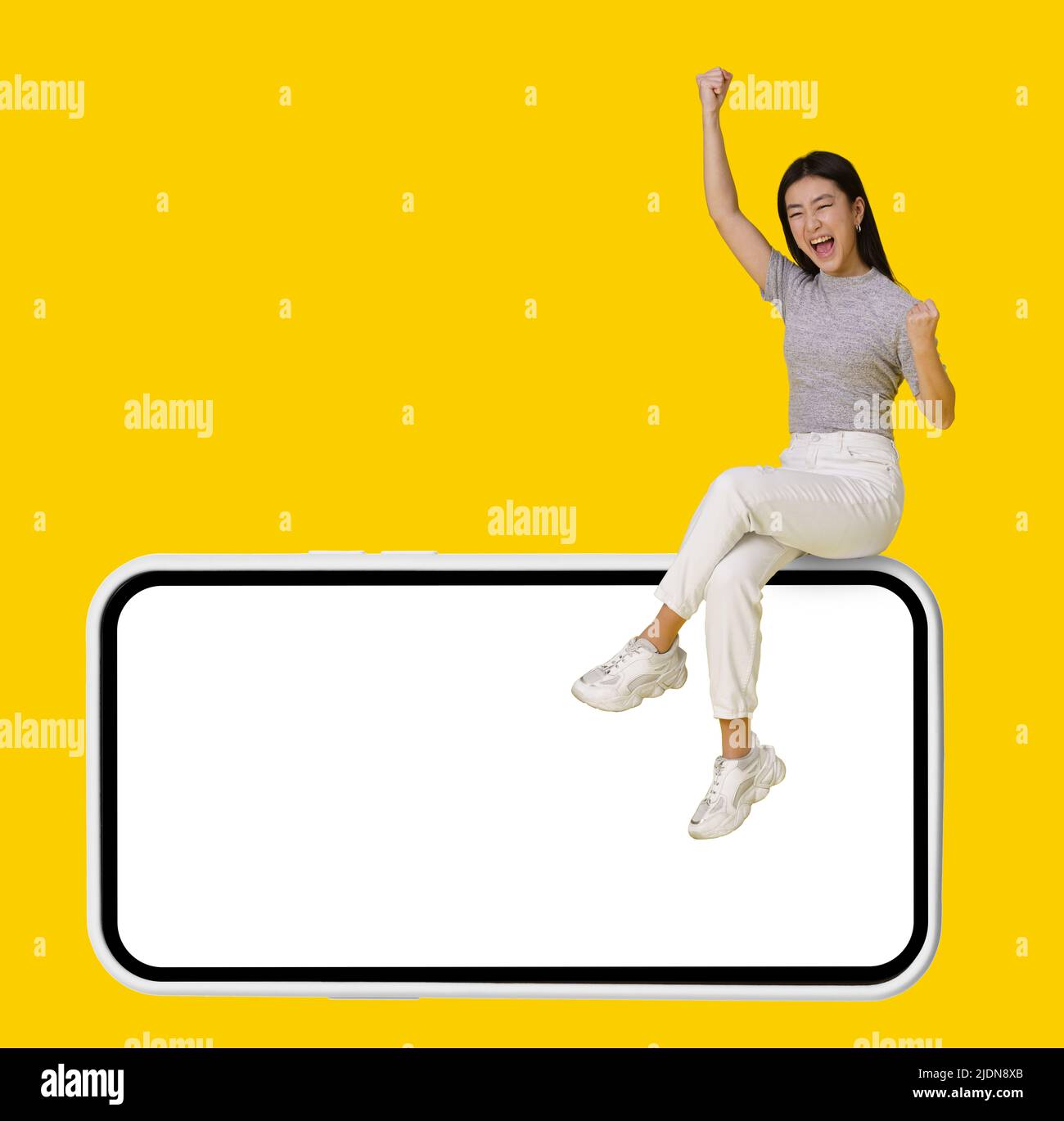 Excited with hands lifted is victory asian girl sitting on giant, huge smartphone with white screen isolated on yellow background. Mock up product placement for mobile app advertisement. Copy space.  Stock Photo