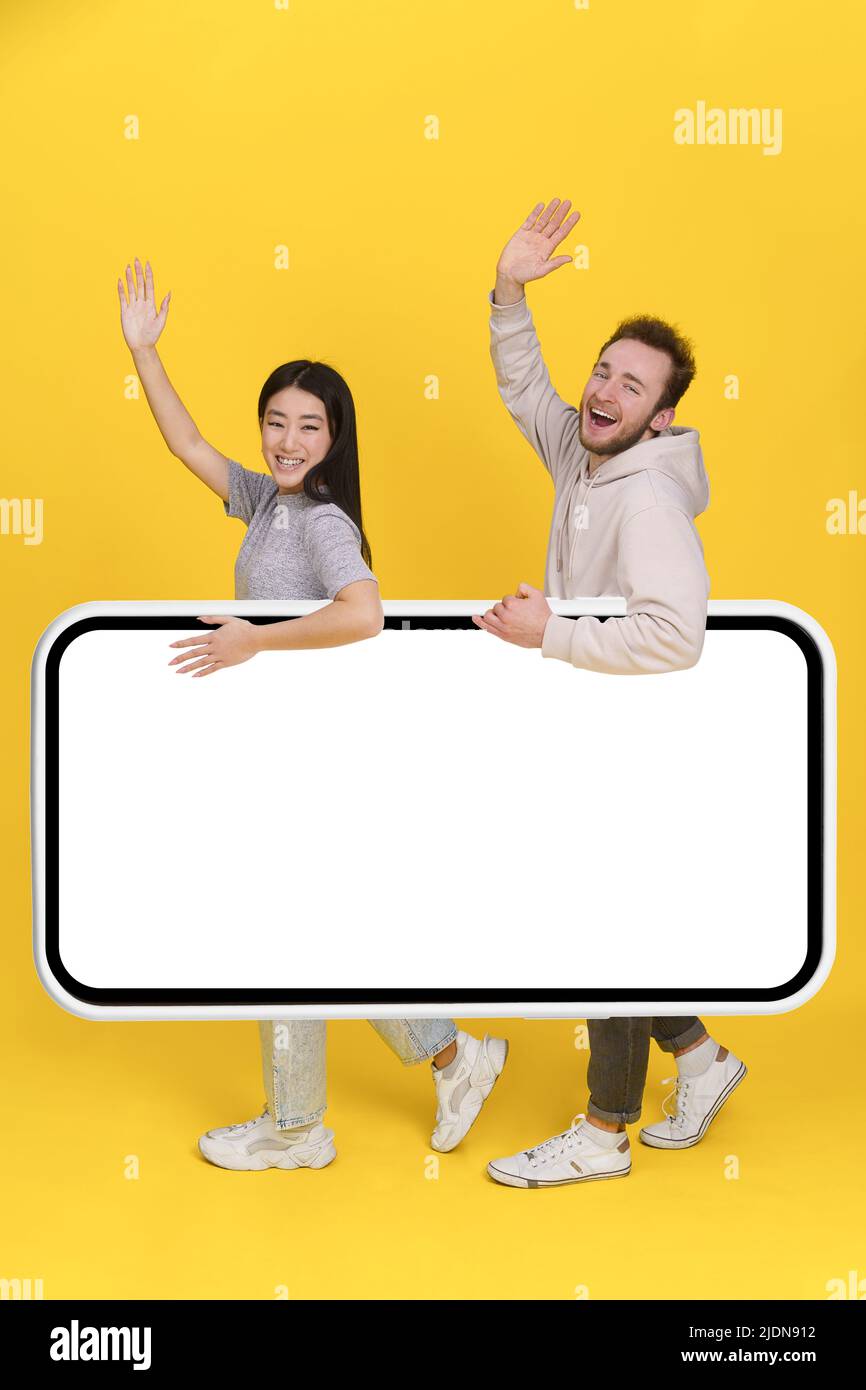 Greeting handsome guy and asian girl holding huge smartphone, digital tablet with white screen, mobile app advertisement and excited smile on camera isolated on yellow background. Product placement.  Stock Photo