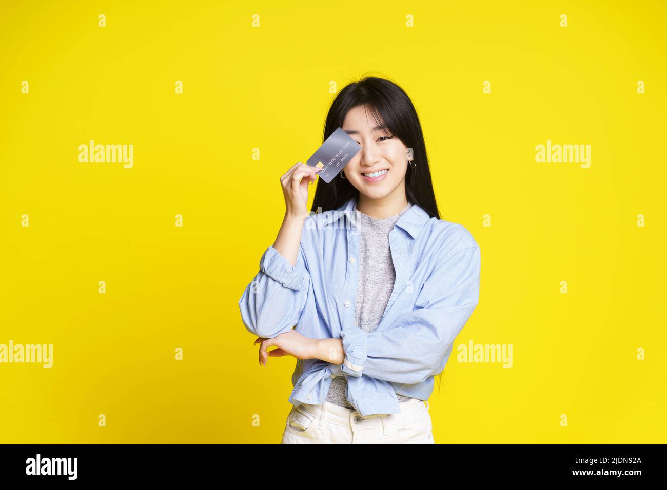 Charming asian girl holding credit, debit card touching her eye. Asian girl in blue shirt holding mockup credit card isolated on yellow background. Shopping concept. Banking concept.  Stock Photo