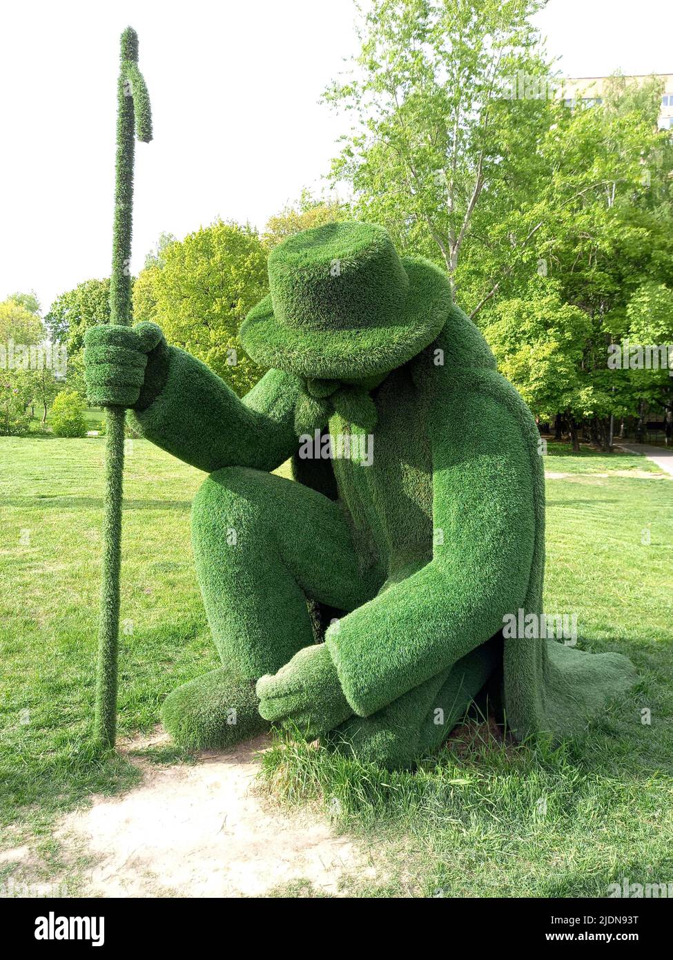 A feeble old man with a stick is trying to get up from the ground. Sculpture made of artificial grass in the city. The concept of helping and caring f Stock Photo