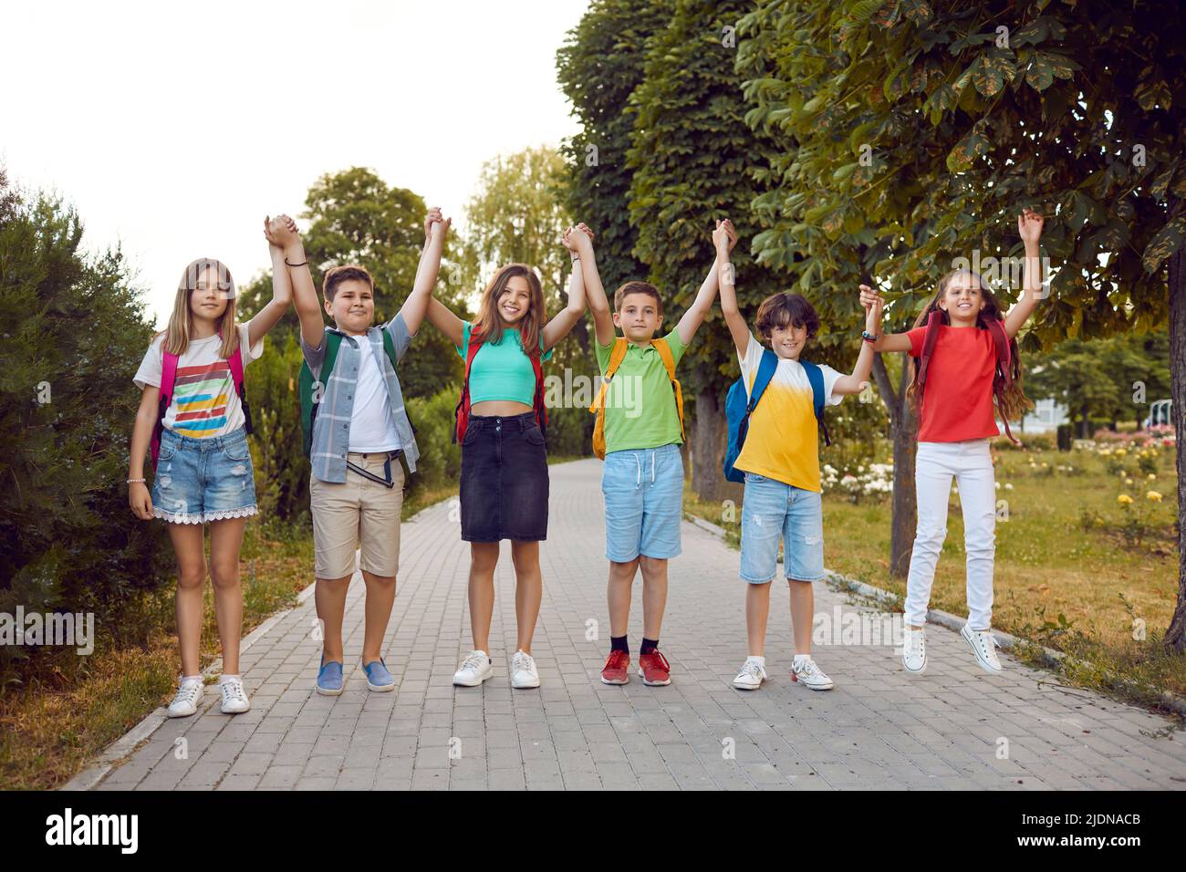 Group of happy school kids with backpacks standing on a park path, holding hands and smiling Stock Photo
