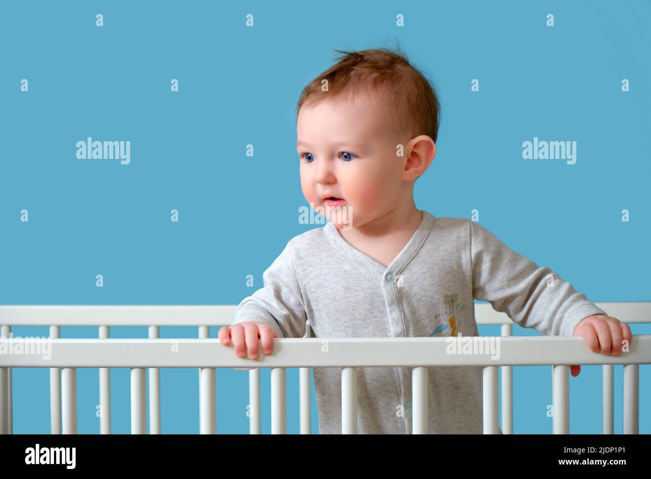Baby boy stands in the crib, studio blue background. Child in white