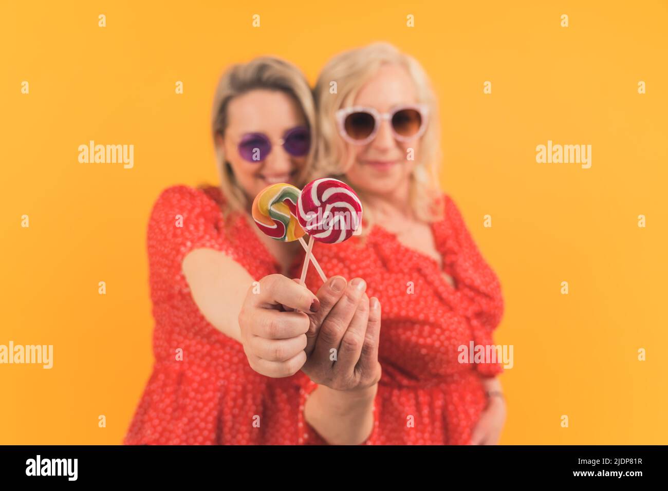 Two blonde excited middle-aged women wearing same red dotted dresses and sunglasses holding colorful lollipops. Yellow background, studio shot. High quality photo Stock Photo