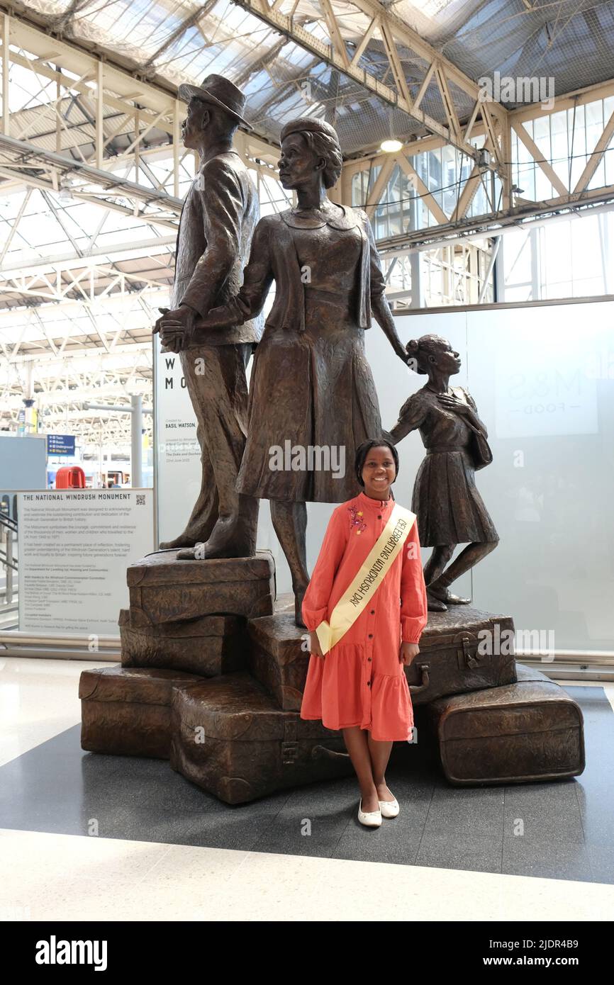 London, UK, 22nd June, 2022. A young girl wearing a windrush day sash poses in front of the National Windrush Monument designed by Jamaican sculptor Basil Watson. It was unveiled on Windrush Day at Waterloo Station, celebrating the Windrush pioneers who first arrived from Caribbean countries at Tilbury Docks in 1948 and up until 1971. The three figures - depicting a man, woman and child symbolises the bond of the Windrush generation. Credit: Eleventh Hour Photography/Alamy Live News Stock Photo