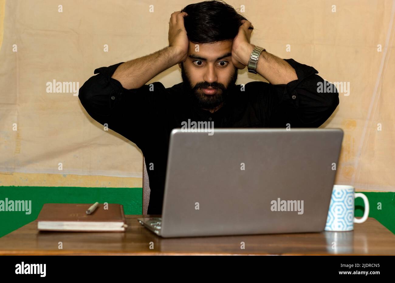 A young Indian man sitting on a chair and giving stressed expression looking at laptop screen. Selective focus on face. Stock Photo