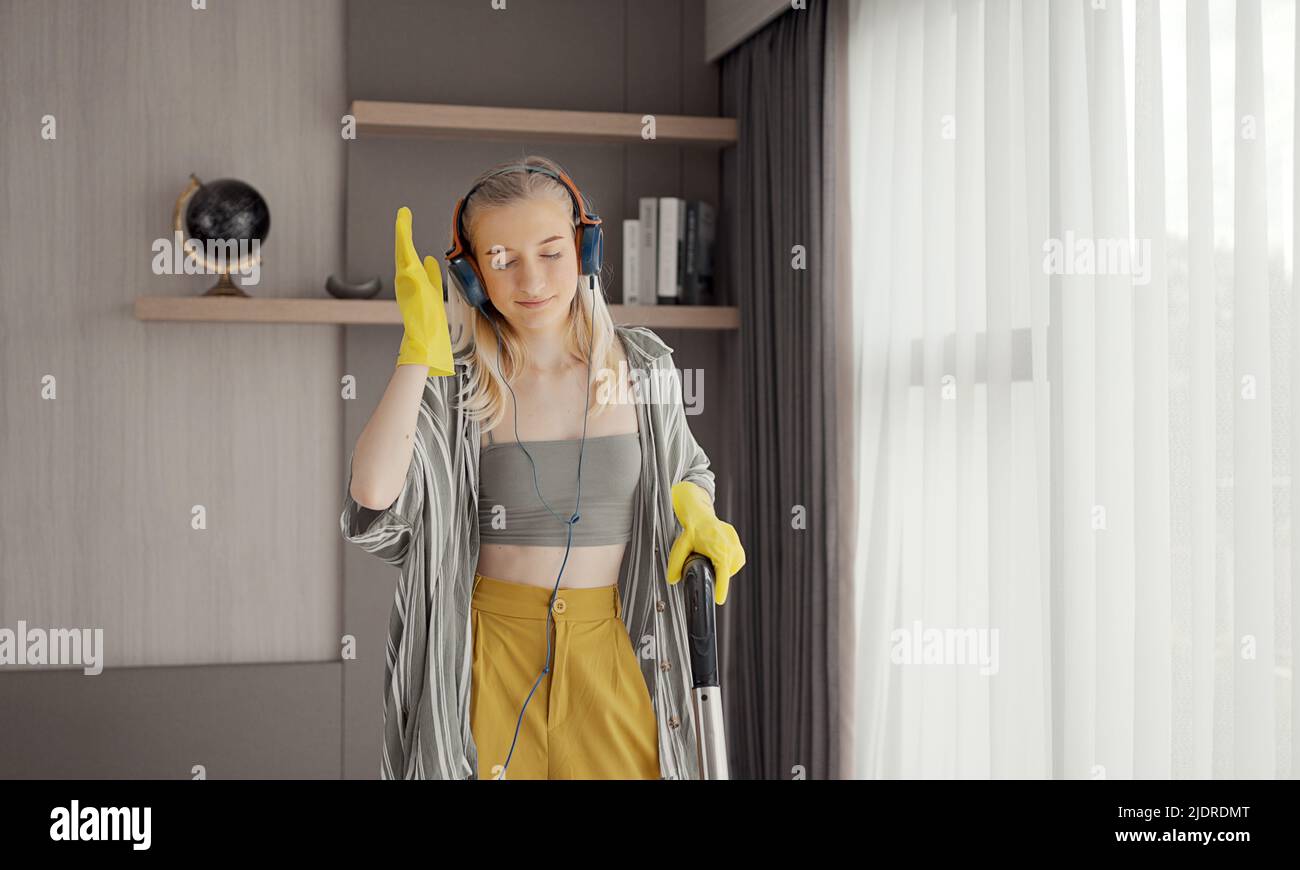 Young girl havig fun while cleaning floor with vacuum cleaner. Happy woman doing housework at home enjoy music wearing earphones. Stock Photo