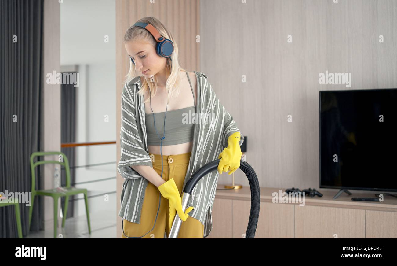 Young girl havig fun while cleaning floor with vacuum cleaner. Happy woman doing housework at home enjoy music wearing earphones. Stock Photo