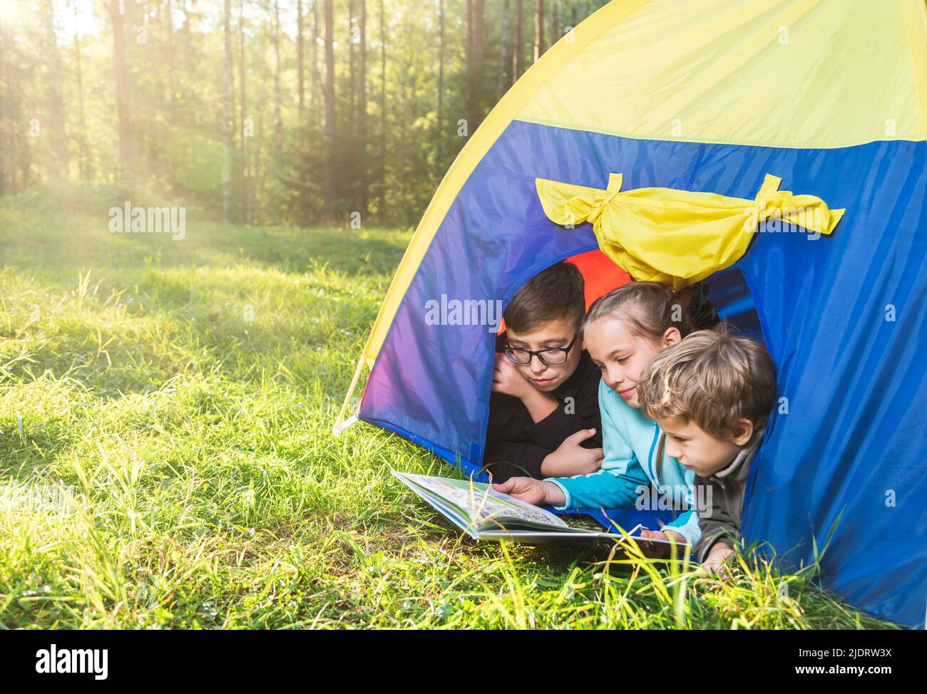 Children reading a book lying together in a tent on a sunny meadow enjoying summer forest camping. The sun shines through the trees. Family summer vac Stock Photo