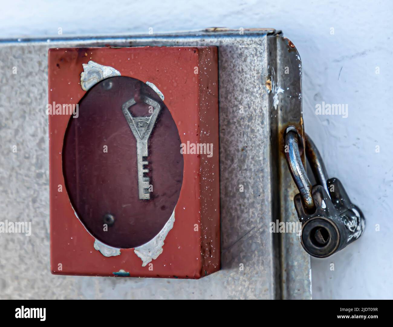A weather-worn red break-glass key holder on a padlocked metal door. Stock Photo
