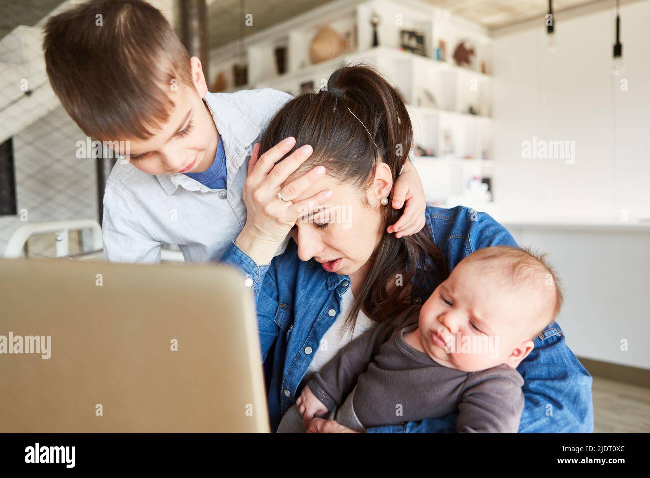 Stressed mother multitasking on pc in home office with baby and big brother Stock Photo
