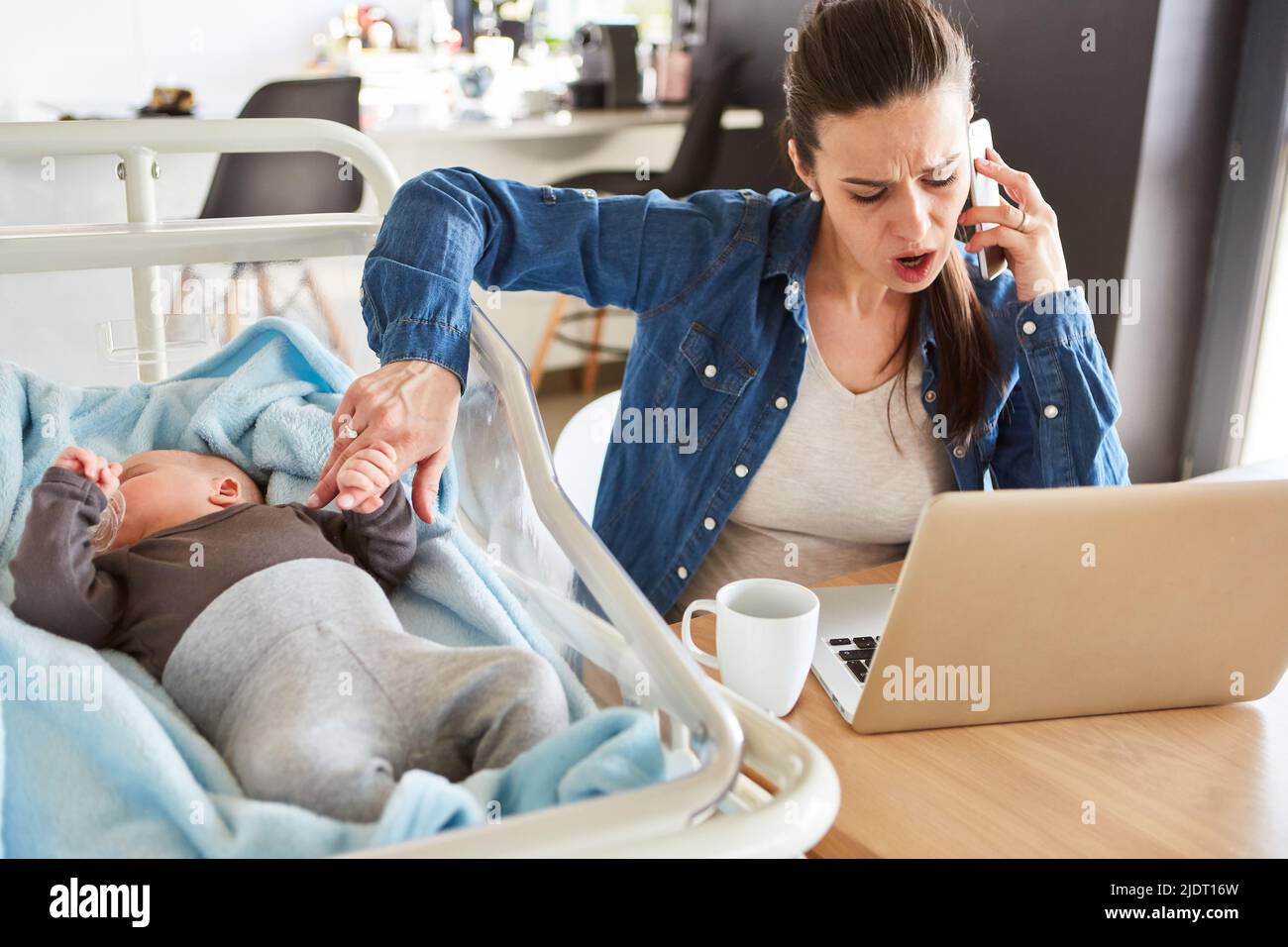 Stressed mother in the home office at the computer looks after her baby and talks on the phone at the same time Stock Photo
