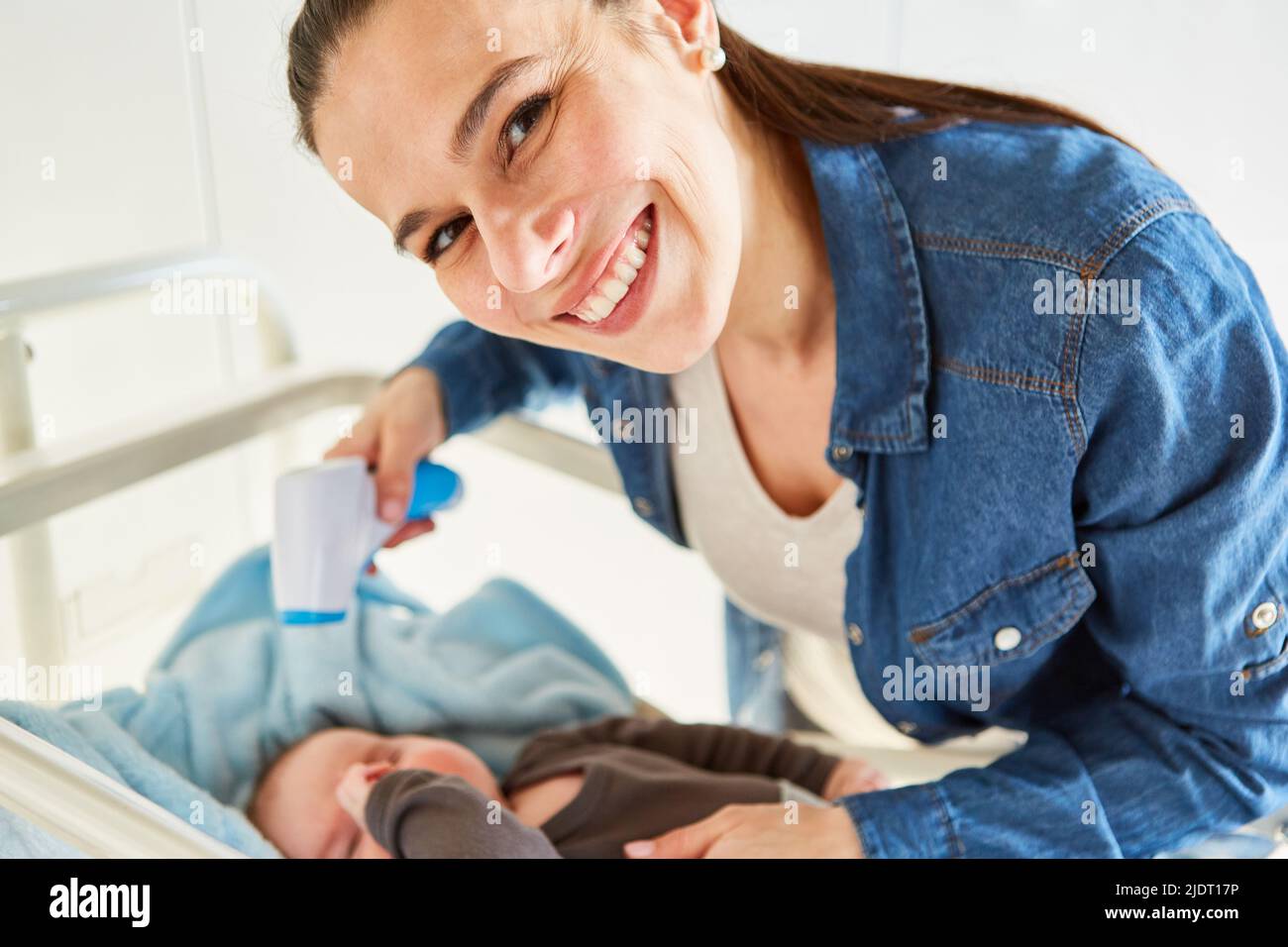 Smiling mother measuring baby's fever in bed with forehead thermometer Stock Photo