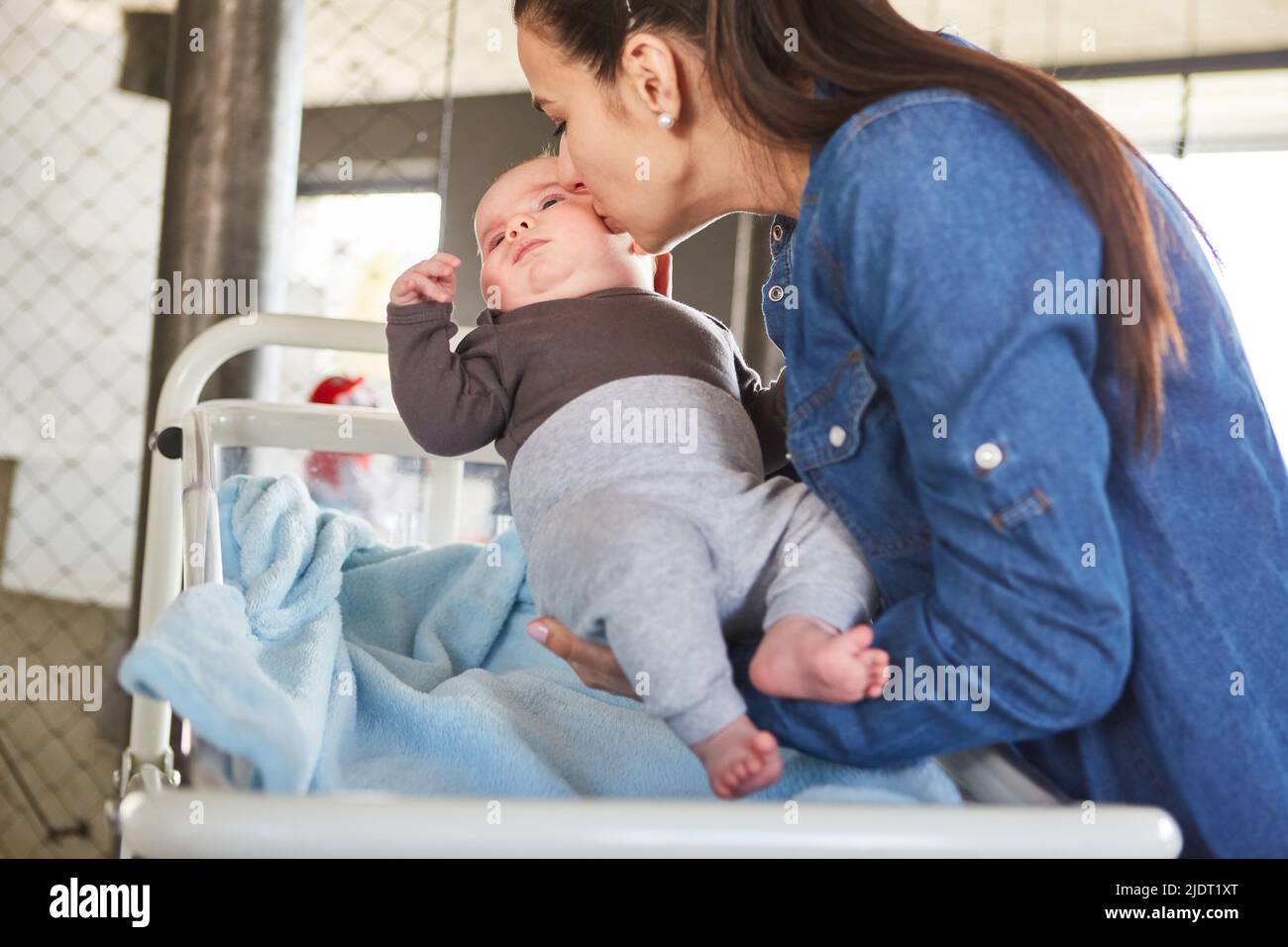 Caring mother carries her baby to the bed and kisses him affectionately on the cheek Stock Photo