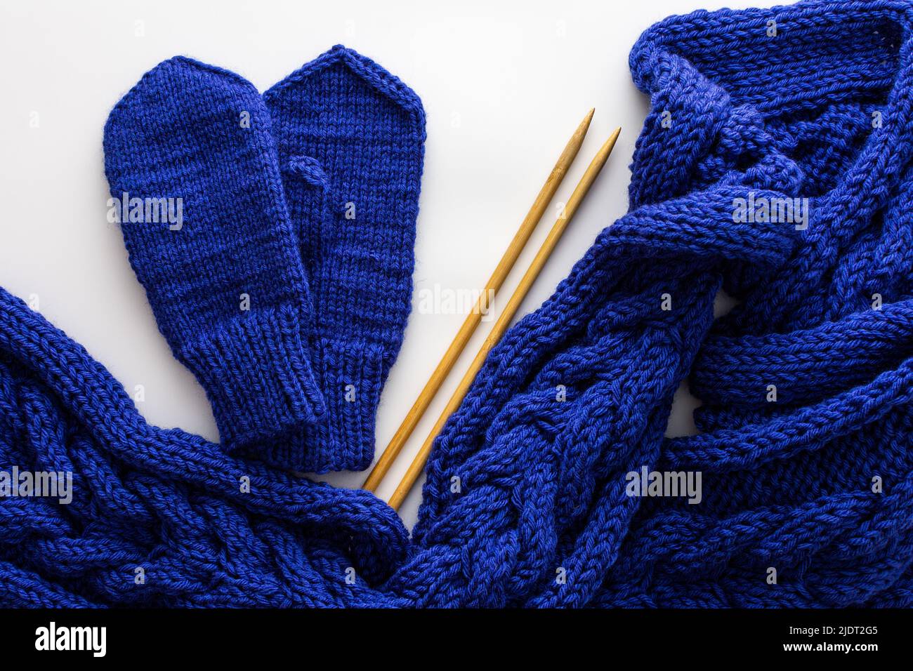 Blue knitted woolen scarf and mittens and knitting needles on a white background, homemade needlework concept, top view Stock Photo