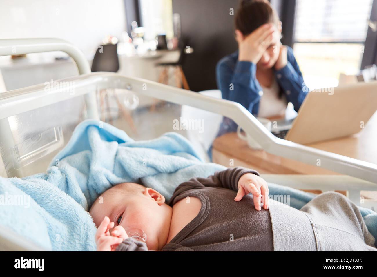 Sleeping baby in cot and exhausted single mother at pc in background Stock Photo