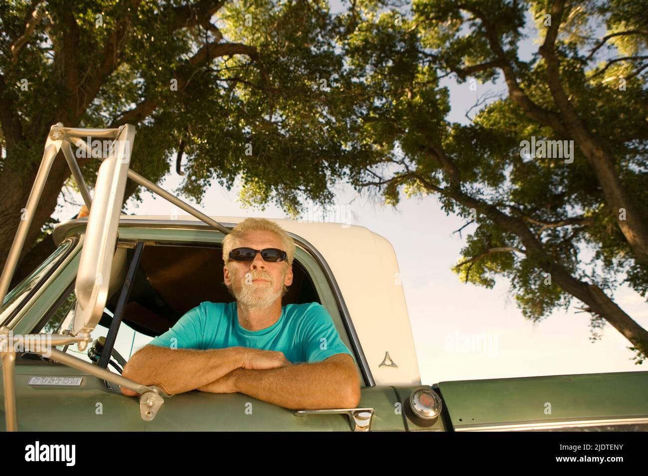 Blonde man ( aged 40-60) with beard wearing sunglasses leaning out the window of a vintage pick up truck parked in a rural area. Stock Photo