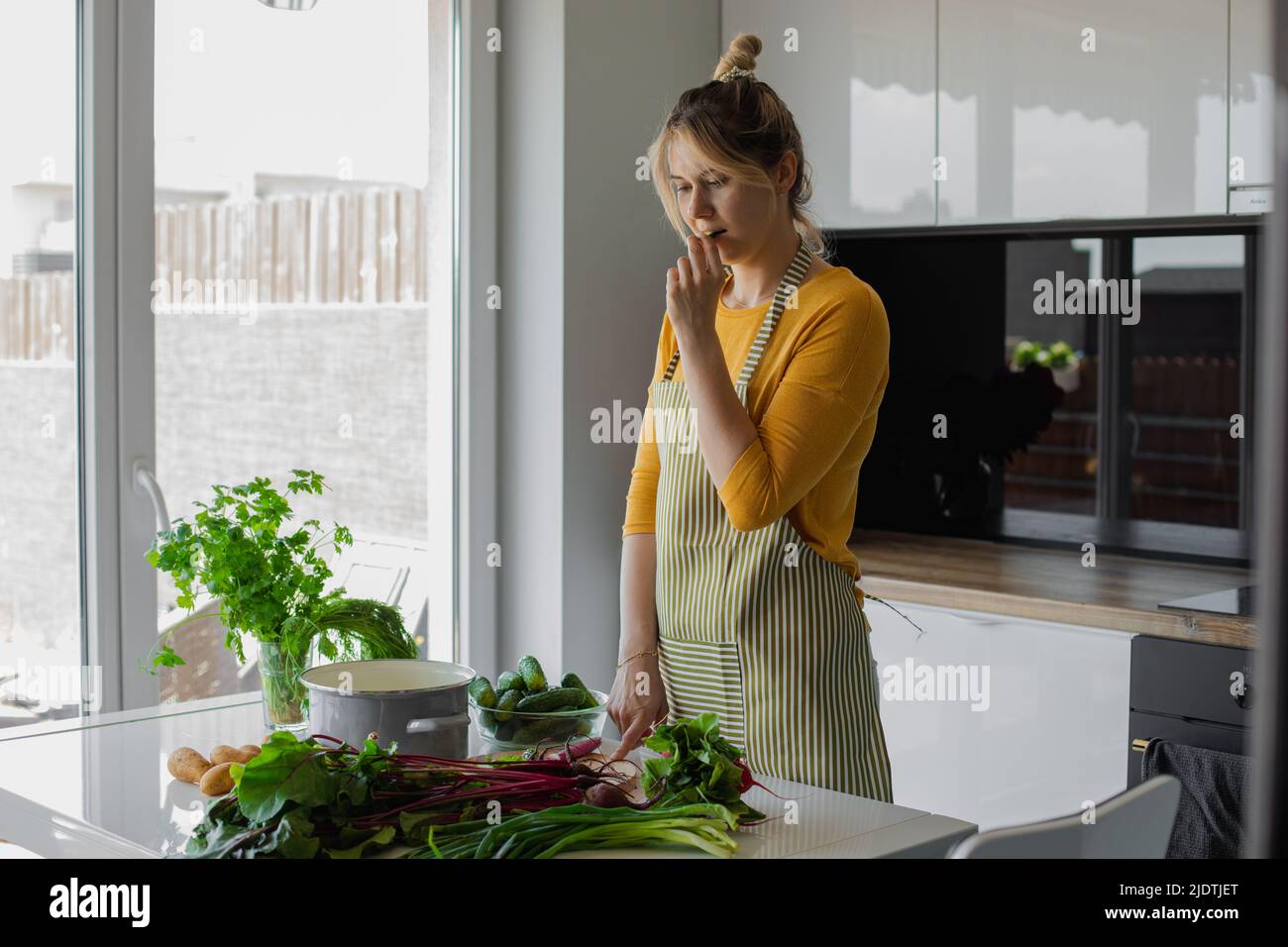 Concentrated woman in apron tasting cucumber, choosing vegetable receipt to cook in the kitchen. Diet, loosing weight Stock Photo