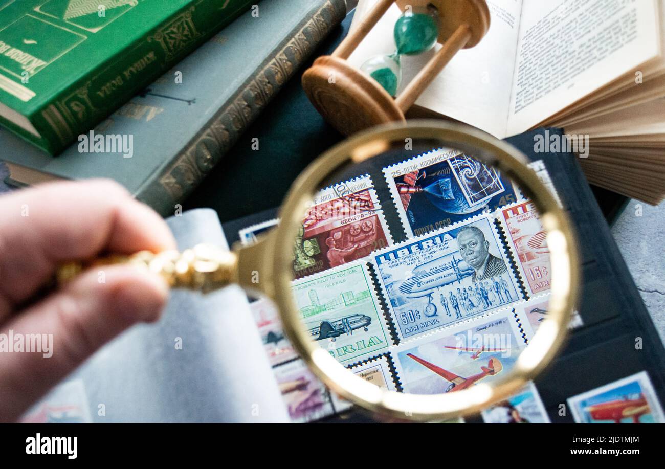 Moscow, Russia, March 2022: Hobby: stamp collecting. Looking at 50 cent stamp of Liberia through a golden magnifying glass. Old books and an hourglass Stock Photo