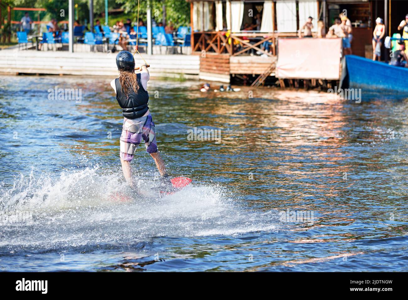 An athlete wakeboarder in a protective helmet and vest goes to the start on the summer bay of the river in front of the stands of spectators. Stock Photo