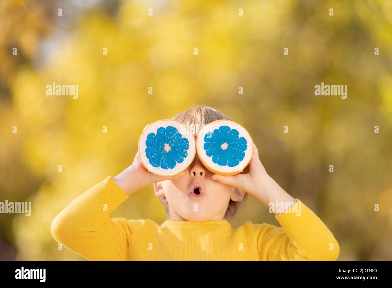 Surprised child holding slices of blue orange fruit like sunglasses. Kid against yellow autumn leaves background. Healthy eating and strange things co Stock Photo