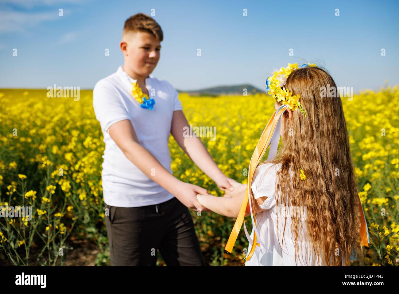Teenagers: brother and sister with Ukrainian wreath with on head, in rapeseed field under blue sky Stock Photo