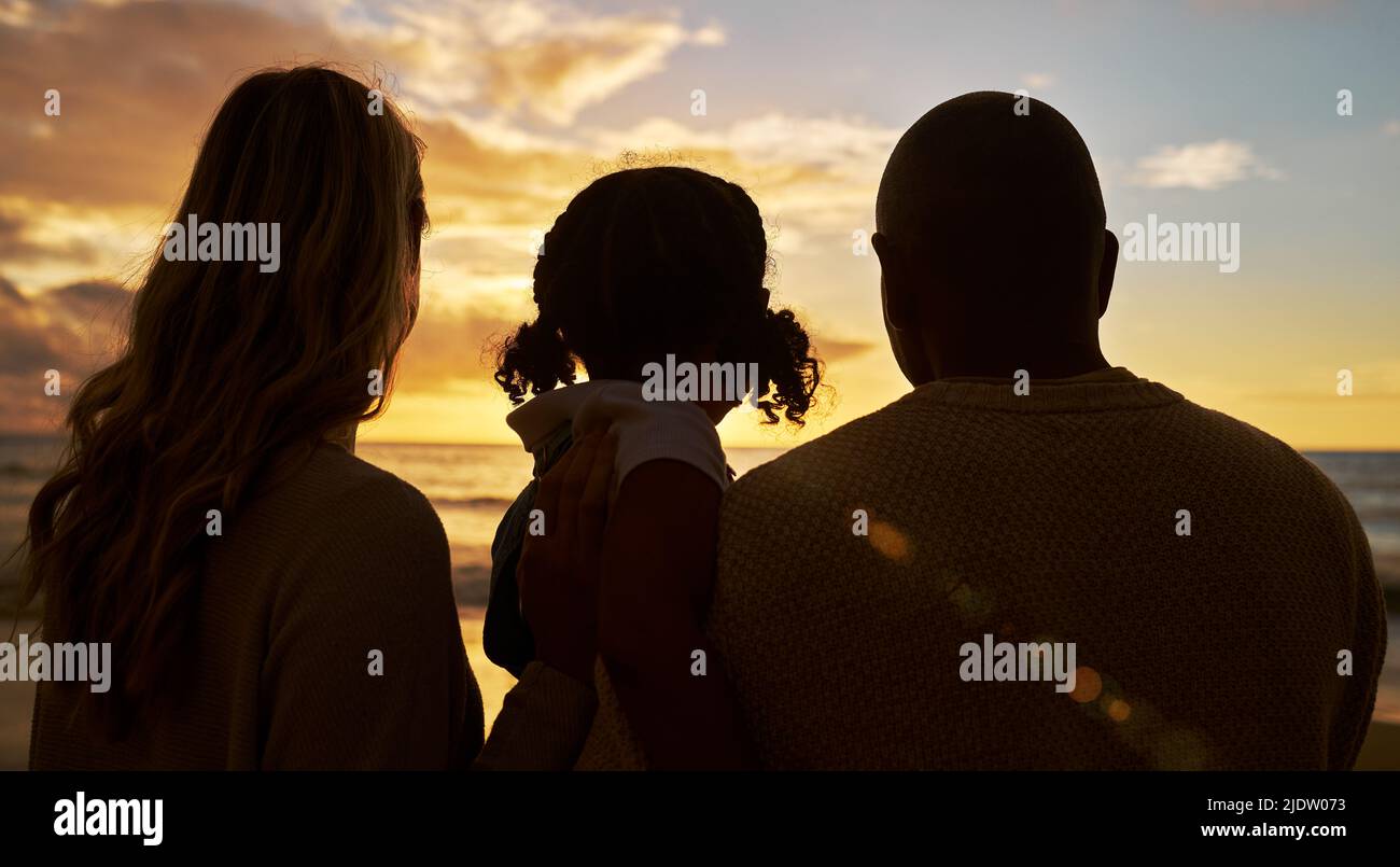 Silhouette closeup of happy family with one child on the beach looking at view at sunset. Two parents and daughter admiring golden sky and calm sea Stock Photo