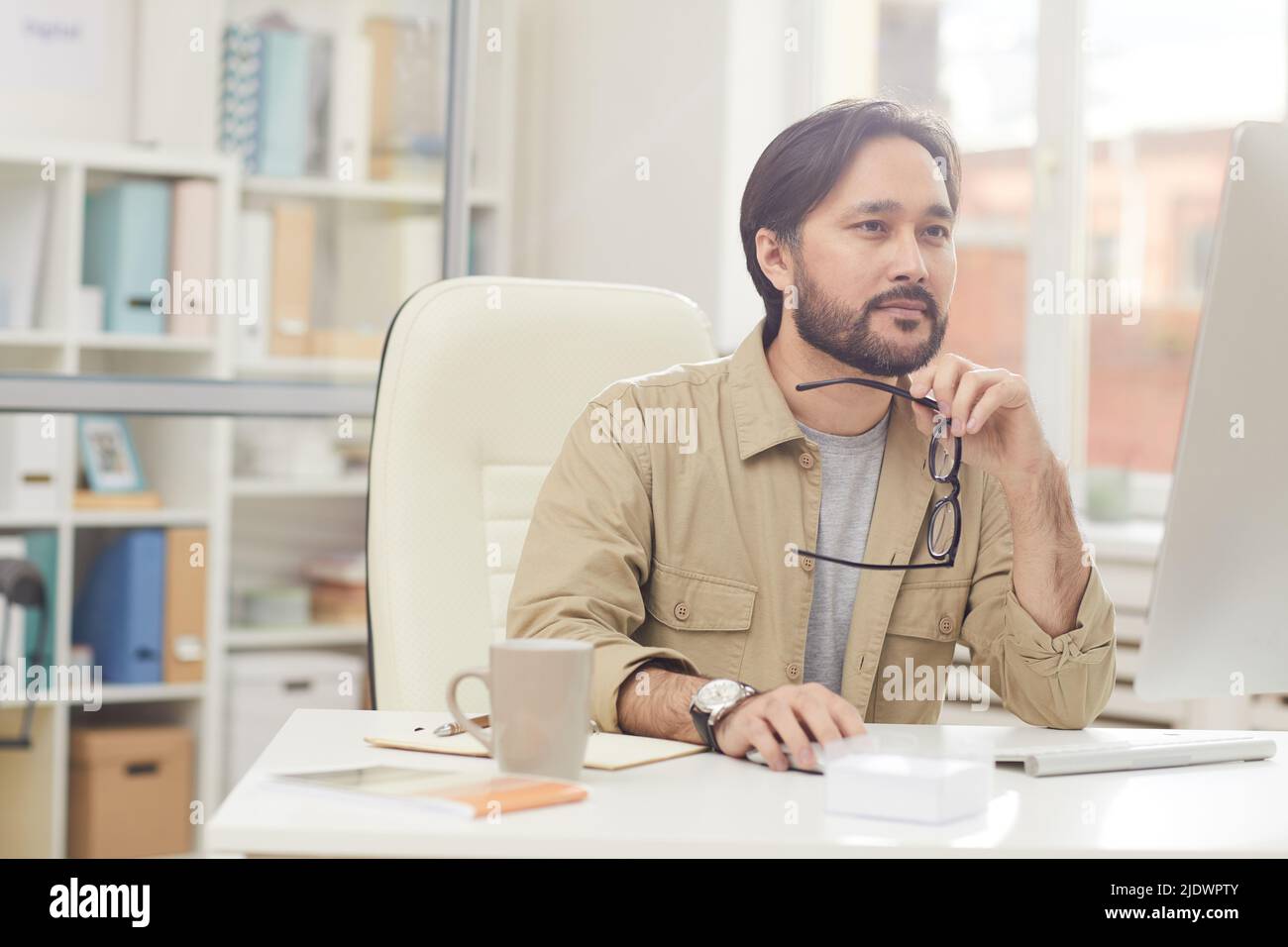 Serious thoughtful young Asian programmer sitting at table and holding eyeglasses while creating design solution Stock Photo