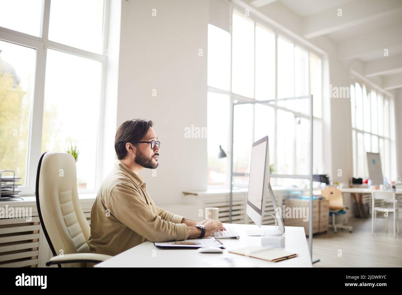 Serious busy young Asian coder in casual shirt and glasses sitting at table and using computer while working with computer language Stock Photo