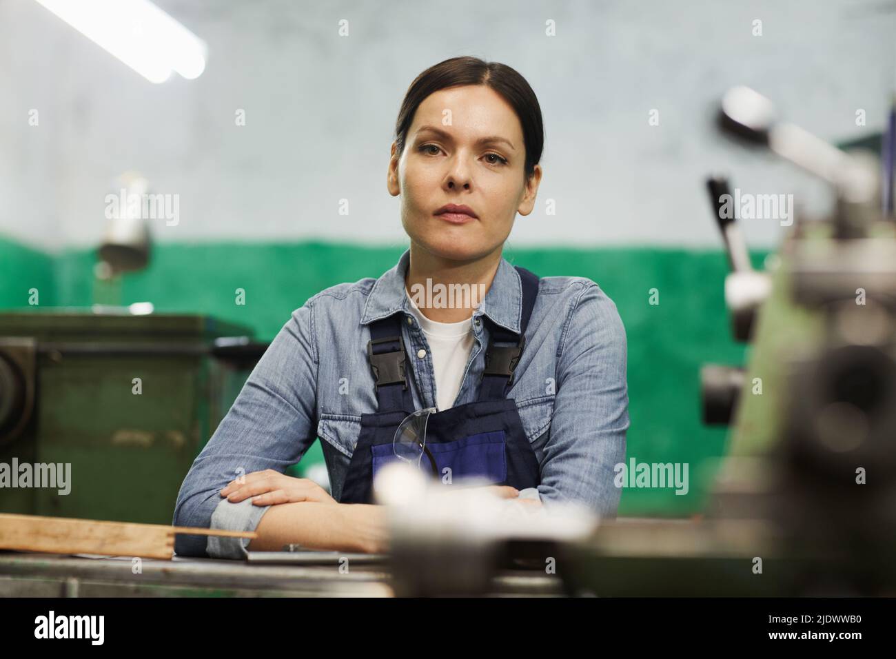 Portrait of serious tenacious lady engineer with brown hair leaning on table at factory workshop Stock Photo