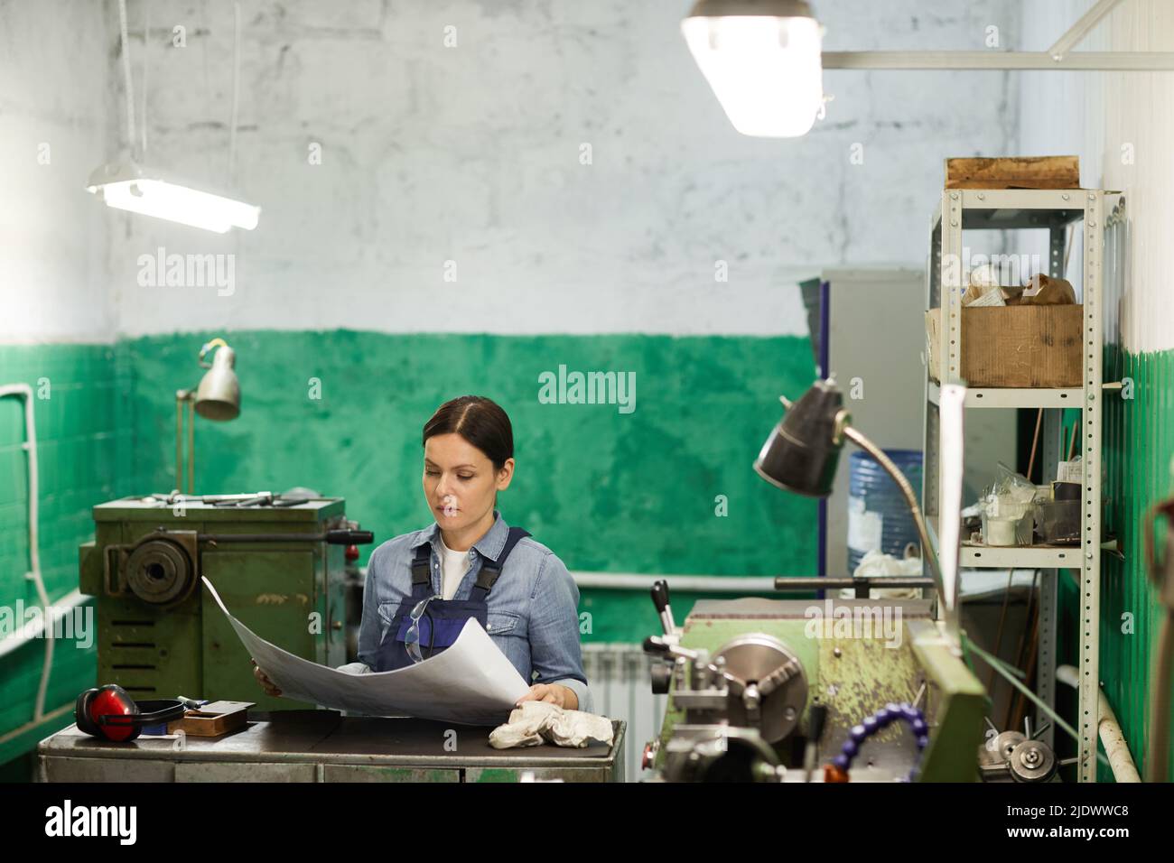 Thoughtful middle-aged female engineer in workwear standing at metal counter and reading technical sketch in industrial shop Stock Photo