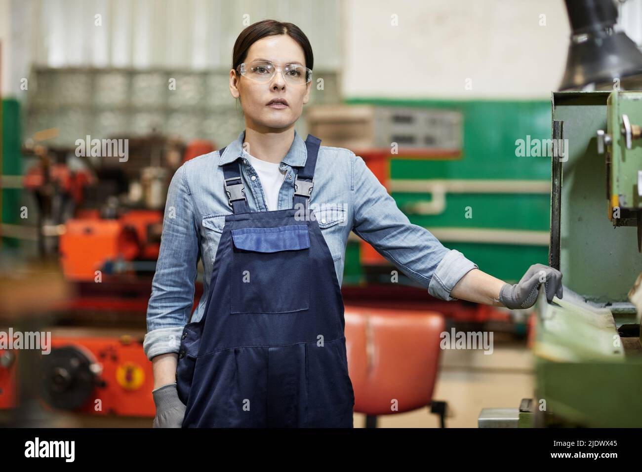 Pensive gritty female manufacturing engineer in blue overalls wearing protective goggles and gloves standing at factory machine Stock Photo