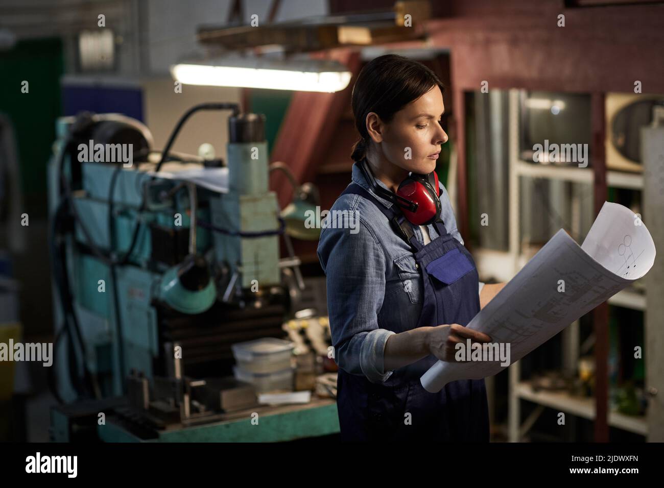 Serious emancipated brunette woman with ear protectors on neck standing by lathe and working with engineering sketch Stock Photo