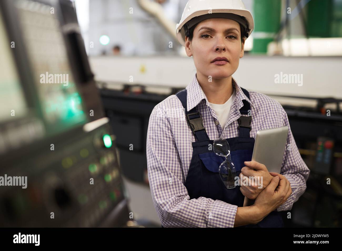Portrait of serious confident female industrial engineer in hardhat and workwear standing at cnc lathe and holding tablet Stock Photo