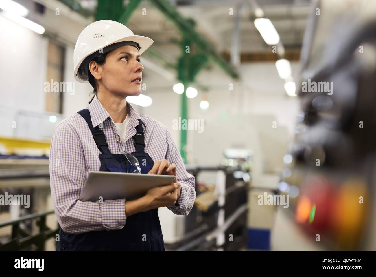 Serious thoughtful female technical engineer in hardhat standing at factory and using tablet for operating cnc machine Stock Photo
