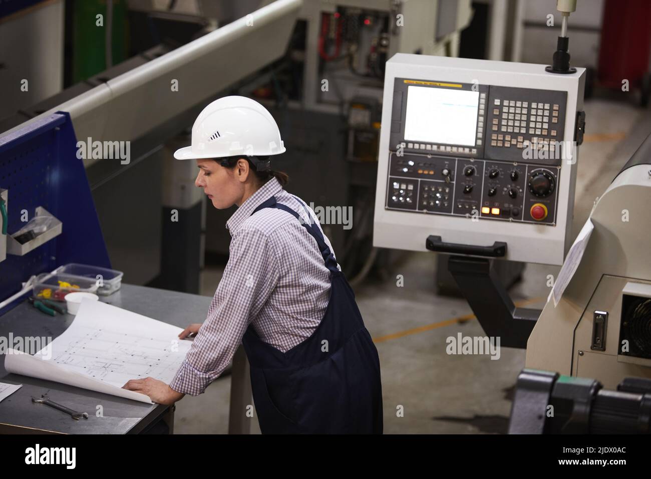 Serious thoughtful female engineer in hardhat standing at desk and studying technical blueprint of metal detail Stock Photo