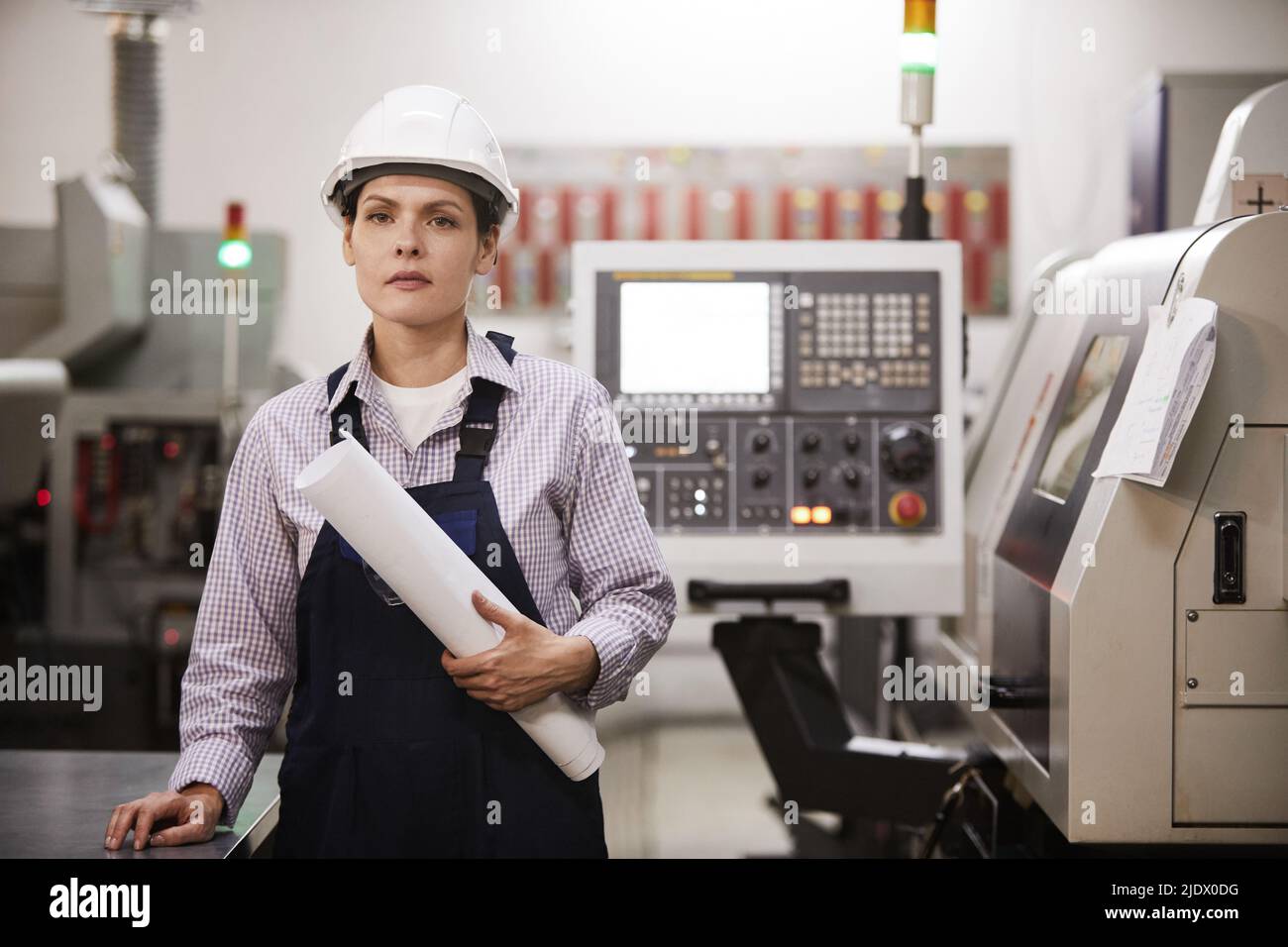 Portrait of serious confident female engineer in hardhat standing at cnc machine and holding rolled blueprint Stock Photo