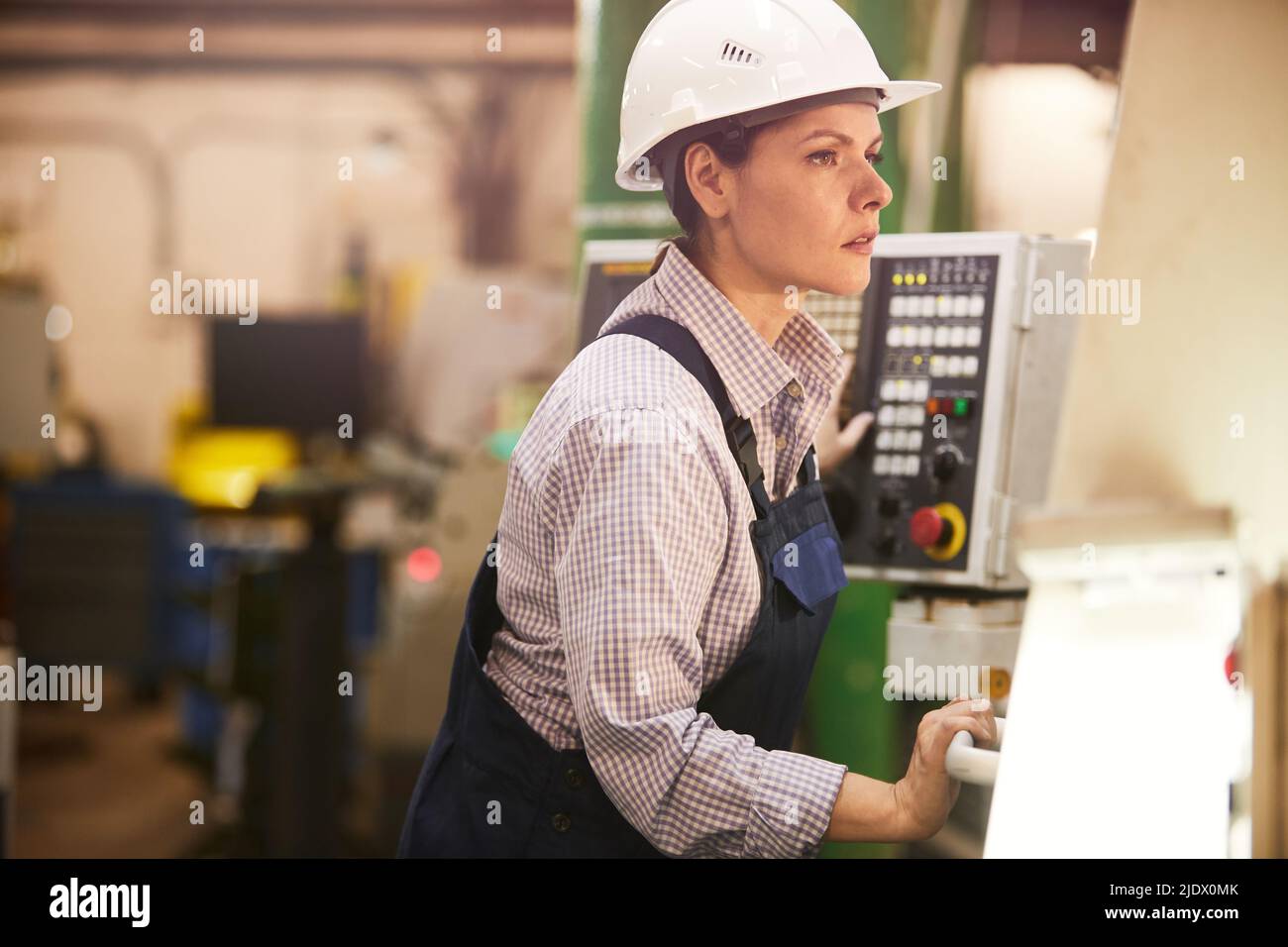 Serious modern female engineer in work helmet standing at factory and controlling work of lathe Stock Photo