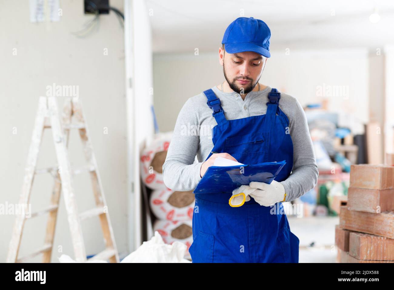 Responsible focused bearded supervisor wearing blue overall taking notes on papers while inspecting construction site Stock Photo