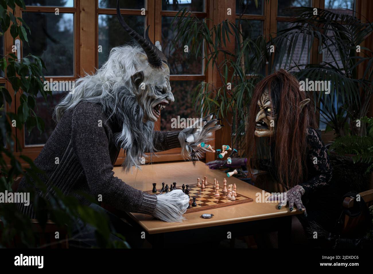 Angry man in spooky costume throwing chess piece on witch sitting at table Stock Photo
