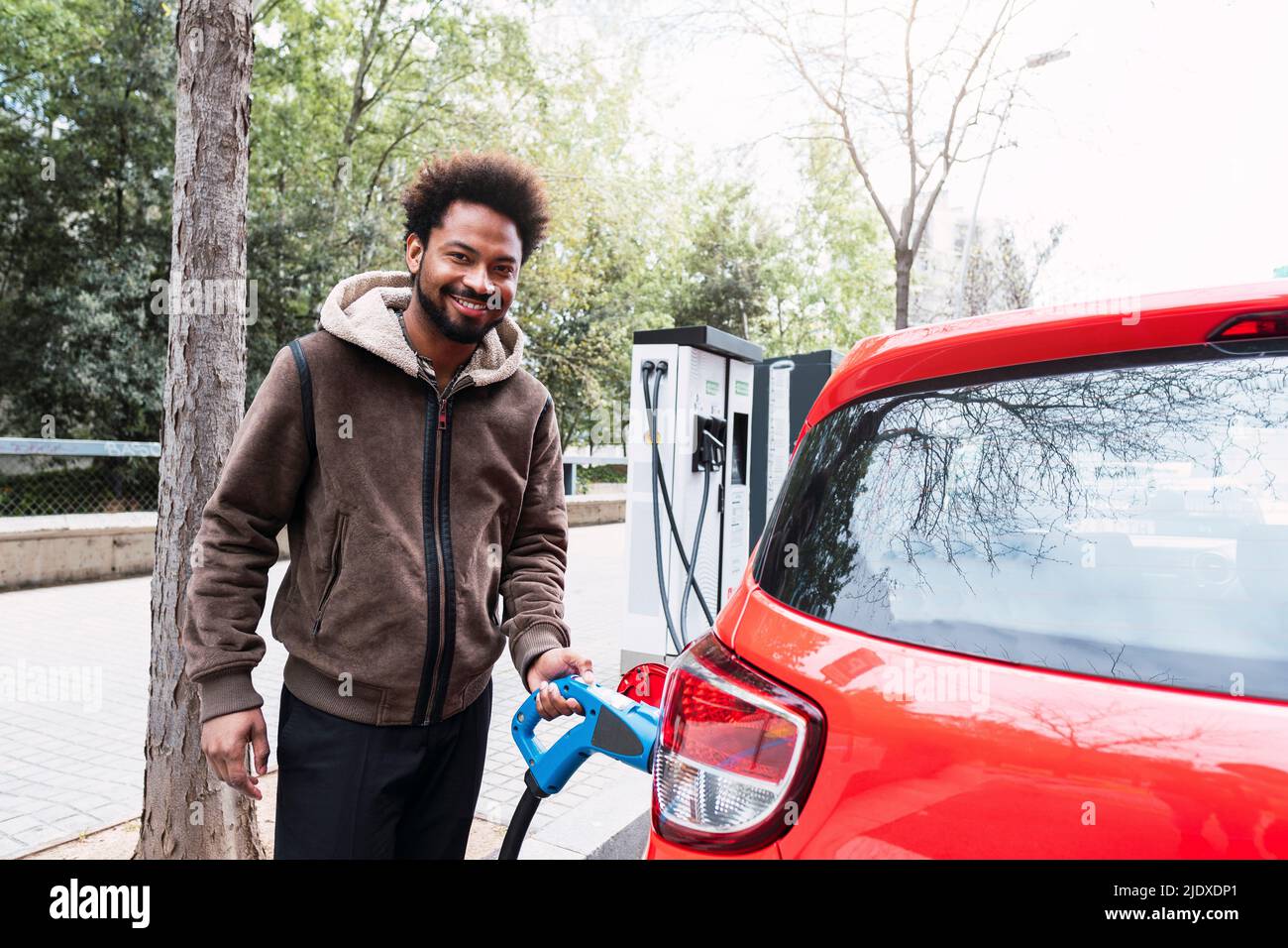 Smiling Afro man charging red electric car at station Stock Photo