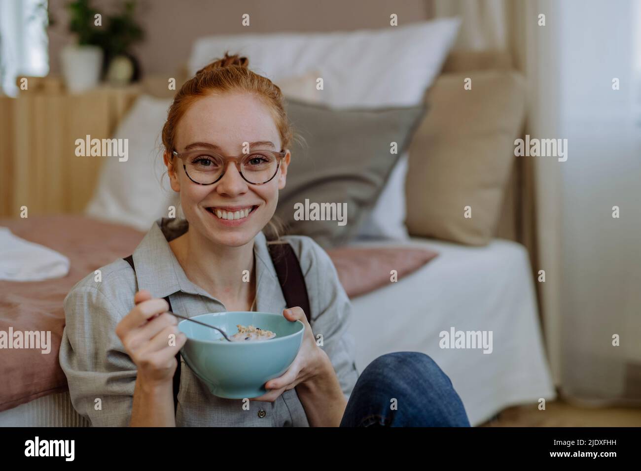 Smiling woman with bowl of food sitting in front of bed at home Stock Photo