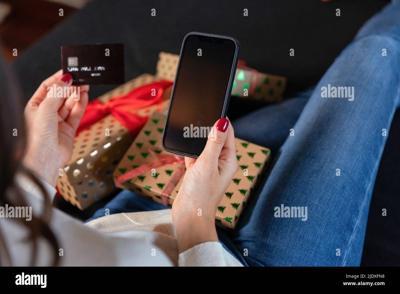 Woman holding credit card doing online shopping through smart phone sitting by Christmas present Stock Photo