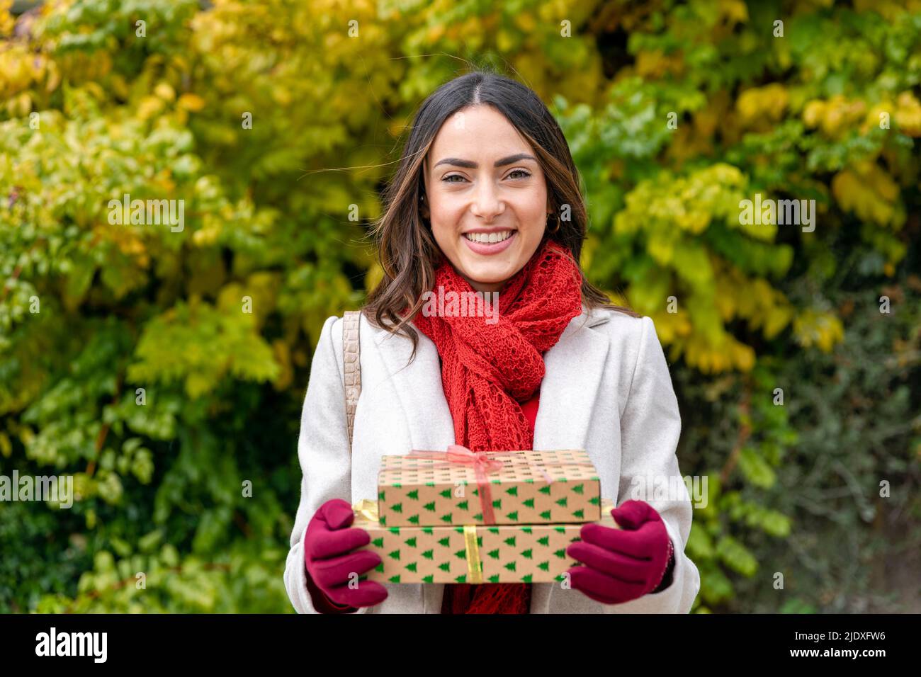 Happy woman wearing red scarf standing with Christmas presents at park Stock Photo
