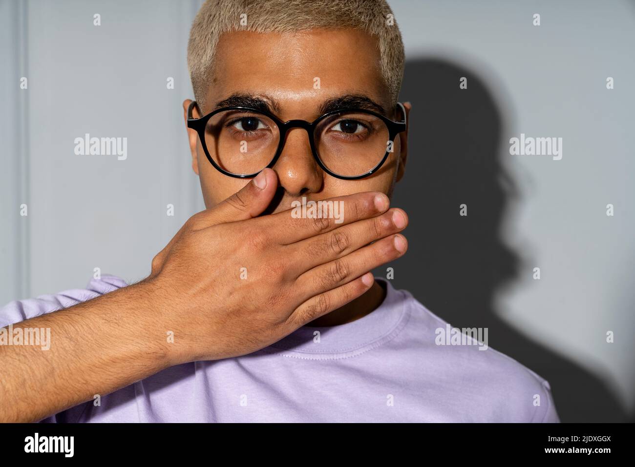 Young man covering mouth with hand in front of wall Stock Photo