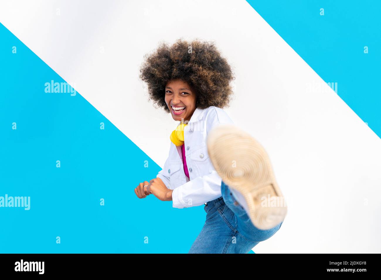 Happy young Afro woman kicking in front of blue and white wall Stock Photo