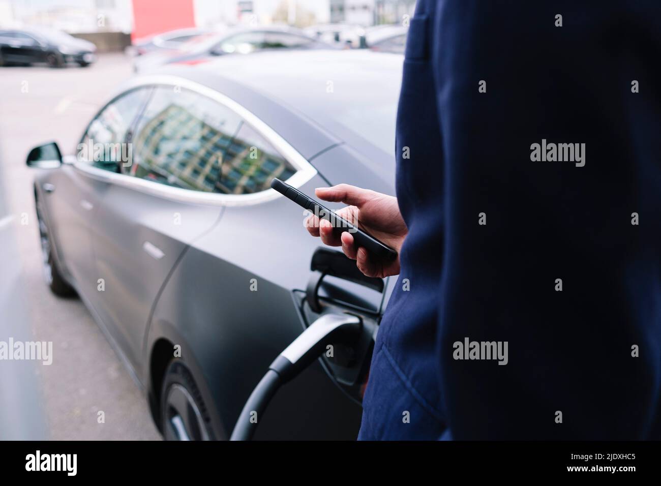 Businessman using smart phone standing by car at electric vehicle charging station Stock Photo