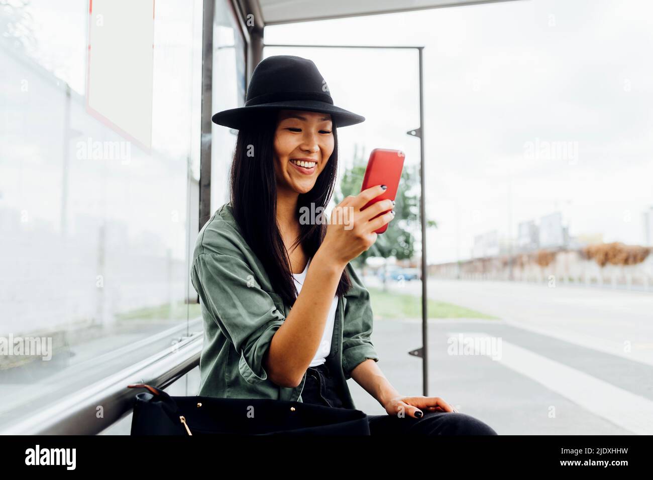 Smiling young woman surfing net through smart phone sitting at bus stop Stock Photo