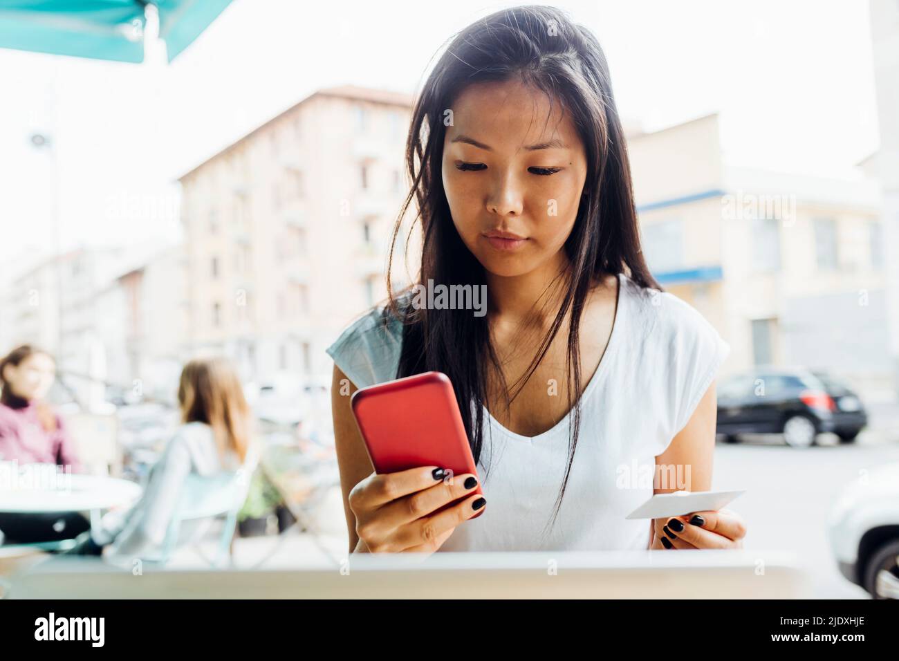 Young woman holding credit card doing online shopping through smart phone sitting at sidewalk cafe Stock Photo