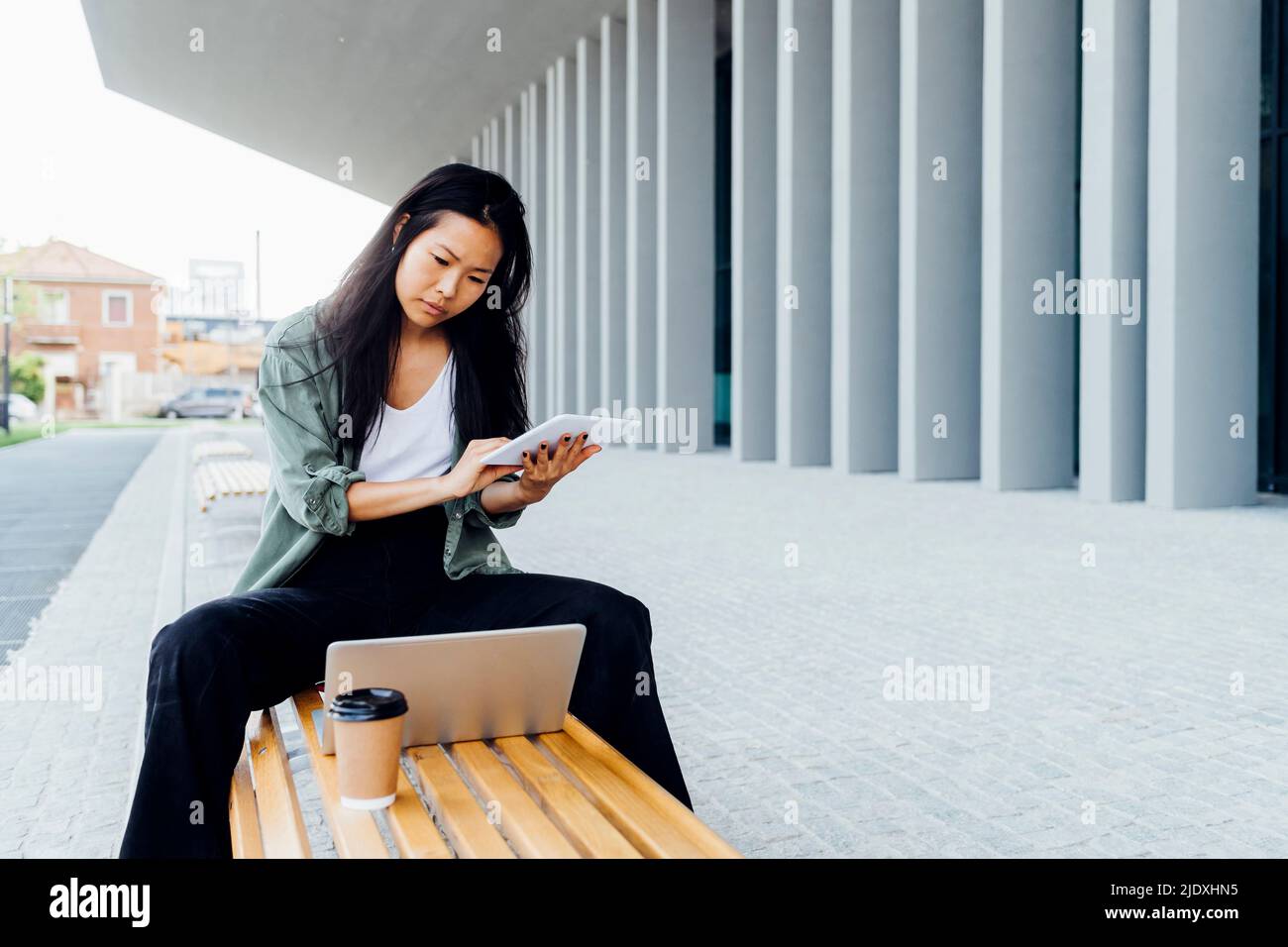 Young freelancer wit tablet PC looking at laptop sitting on bench Stock Photo