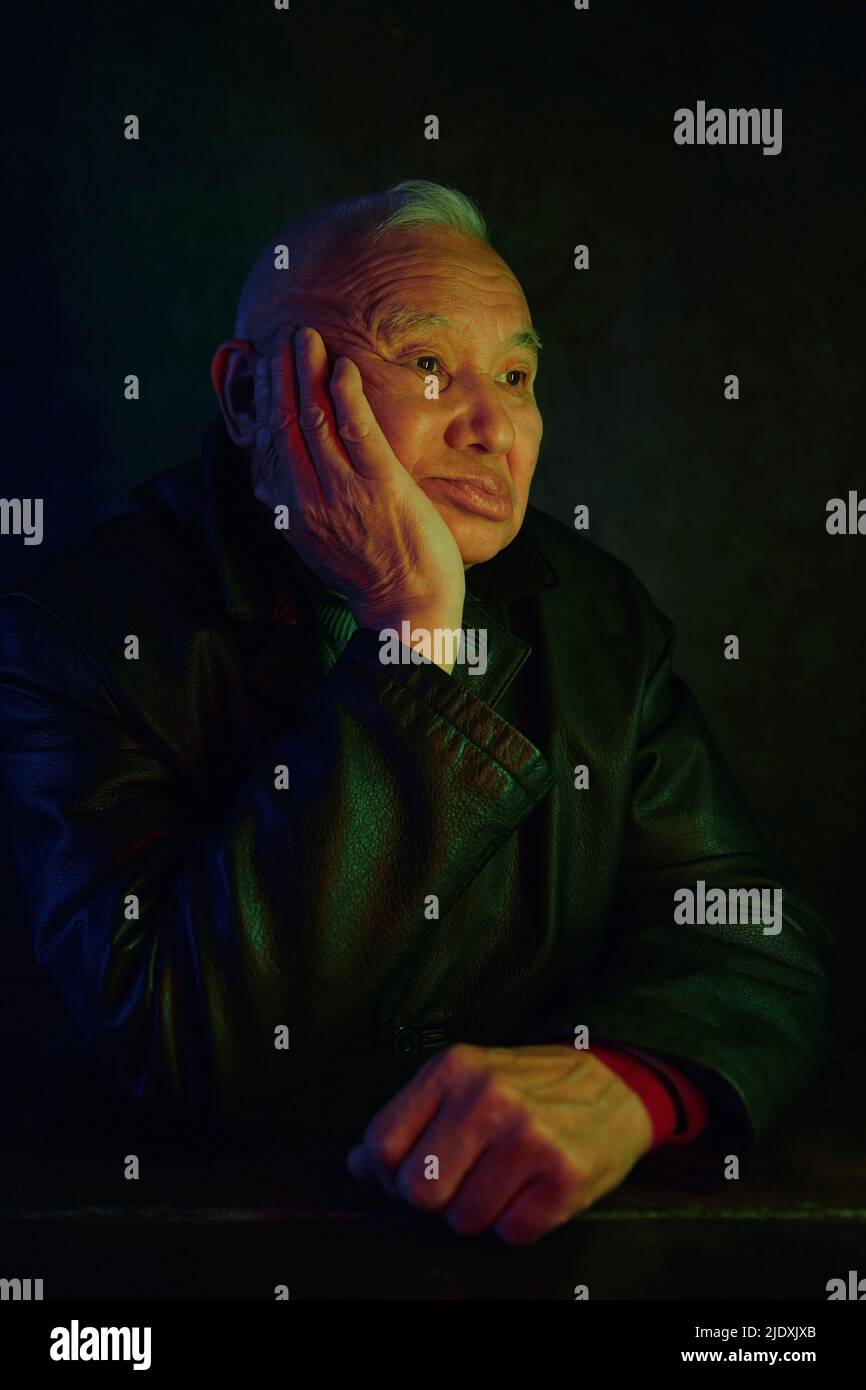 Pensive senior man sitting with hand on chin against black background Stock Photo